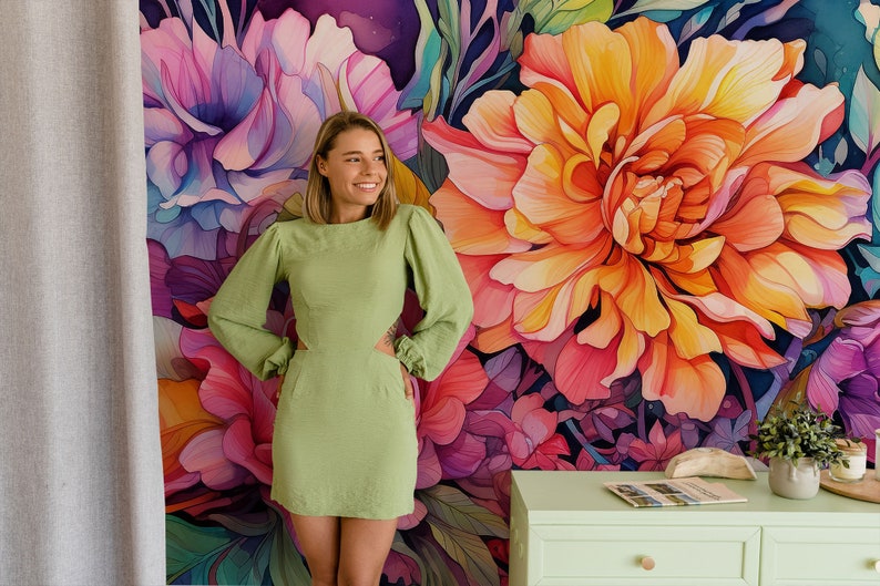 May include: A woman in a light green dress stands in front of a floral mural. The mural features large, colorful flowers in shades of pink, purple, and orange. A light green cabinet with a few items on top is visible.