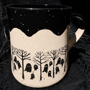 May include: A ceramic mug with a matte beige base and a glossy black rim. The lower section features a black screen-printed design of multiple small ghost figures interspersed among bare, leafless trees against a speckled, light-colored background.