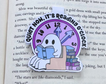 Könnte beinhalten: Ein weißer Geist liest ein Buch mit einem lila Zifferblatt hinter sich. Das Zifferblatt zeigt "Quiet Now, It's Reading O'Clock". Der Geist sitzt auf einem Stuhl mit einem Stapel Bücher hinter sich. Das Zifferblatt ist von einem lila Kreis mit weißem Rand umgeben.