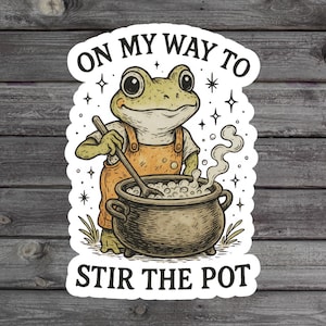 May include: A sticker featuring a cartoon frog wearing an orange apron, stirring a pot. The text reads "ON MY WAY TO STIR THE POT" in a whimsical font, surrounded by stars. The frog is light green and the pot is dark brown.