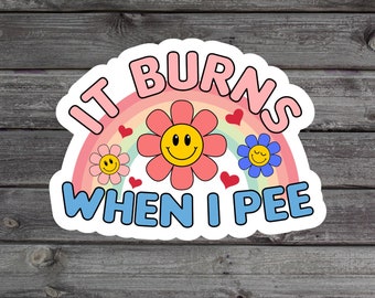 May include: A white sticker with a rainbow design and the text "It burns when I pee". The rainbow has three pink flowers with yellow smiley faces and two hearts.