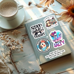 May include: A collection of colorful stickers with humorous phrases and illustrations, including raccoons wearing sunglasses, a chicken, and a skeleton. The stickers are on a gray book, with a cup of coffee and flowers in the background.