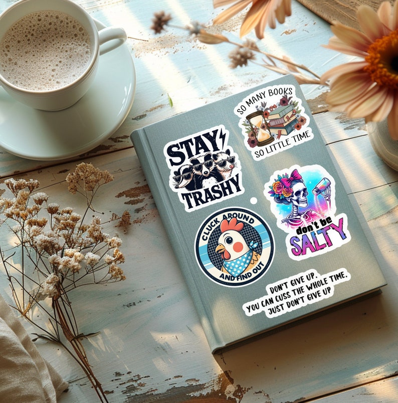 May include: A collection of colorful stickers with humorous phrases and illustrations, including raccoons wearing sunglasses, a chicken, and a skeleton. The stickers are on a gray book, with a cup of coffee and flowers in the background.