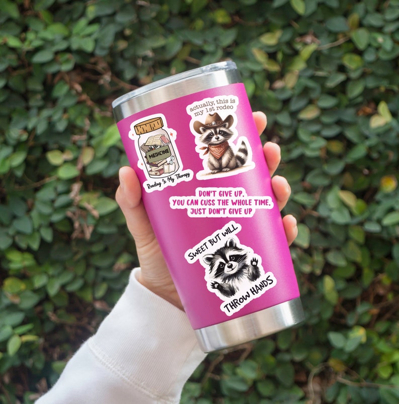 May include: A pink stainless steel tumbler adorned with several stickers. The stickers feature raccoon illustrations, text that reads "Reading is My Therapy," and phrases like "Don't Give Up" and "Sweet But Will Throw Hands."