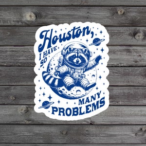 May include: A blue and white sticker with a raccoon wearing an astronaut suit and sitting on the moon. The text reads "Houston, I have so many problems".