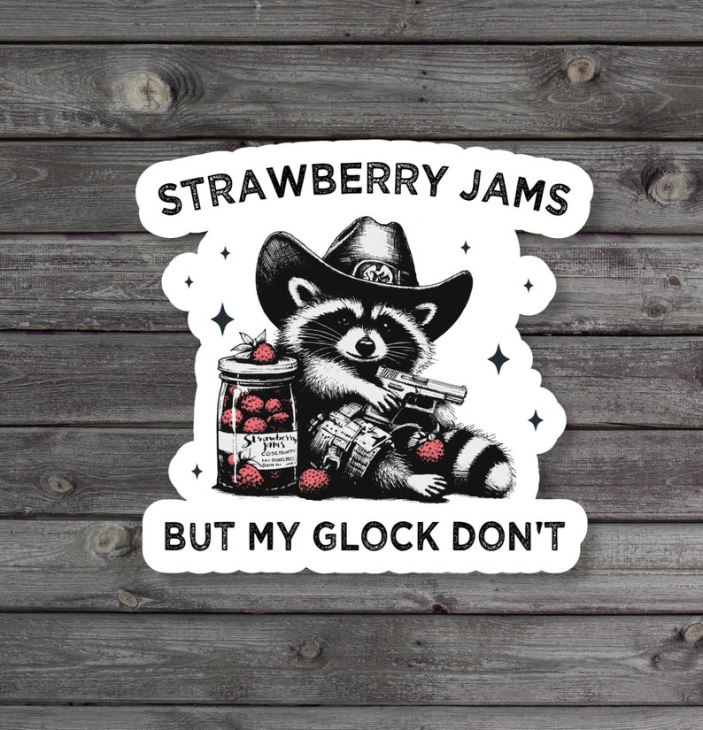 May include: A white sticker with a black and white illustration of a raccoon wearing a cowboy hat and holding a gun. The raccoon is sitting next to a jar of strawberry jam. The text "Strawberry Jams But My Glock Don't" is printed on the sticker.