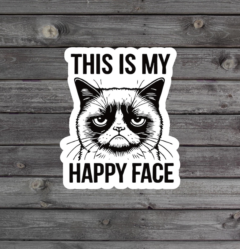 May include: A white sticker with a black and white illustration of a grumpy cat with the text "This is my happy face" on a wood background.