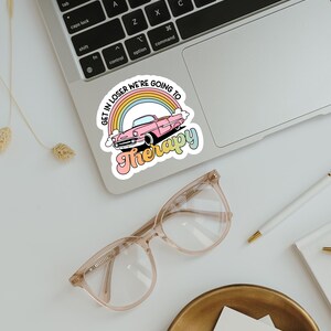 Pu&ograve; includere: Un adesivo con la scritta "GET IN LOSER WE'RE GOING TO Therapy" in un font retr&ograve;. L'adesivo presenta un'auto rosa classica davanti a un arcobaleno. L'adesivo &egrave; su un laptop.