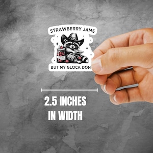 May include: A black and white illustration of a raccoon wearing a cowboy hat and holding a jar of strawberry jam and a gun. The text reads "Strawberry Jams But My Glock Don". The sticker is 2.5 inches wide.