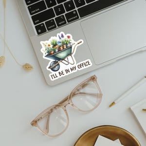 May include: A sticker featuring a watercolor illustration of a wheelbarrow filled with potted plants and flowers. The sticker has the text "I'LL BE IN MY OFFICE" in a handwritten font. The sticker is placed on a laptop.