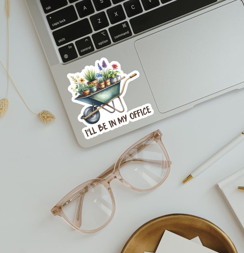 May include: A sticker featuring a watercolor illustration of a wheelbarrow filled with potted plants and flowers. The sticker has the text "I'LL BE IN MY OFFICE" in a handwritten font. The sticker is placed on a laptop.