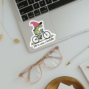 May include: A sticker featuring a green frog riding a bicycle and wearing a wizard hat. The sticker has the text "Off to cause a kerfuffle". The sticker is on a laptop.