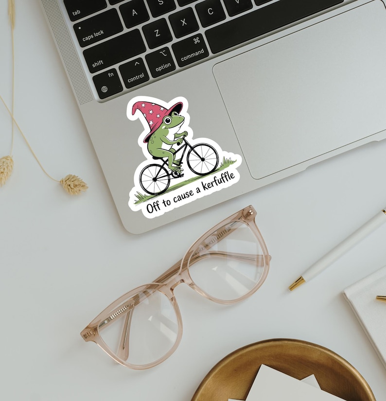 May include: A sticker featuring a green frog riding a bicycle and wearing a wizard hat. The sticker has the text "Off to cause a kerfuffle". The sticker is on a laptop.