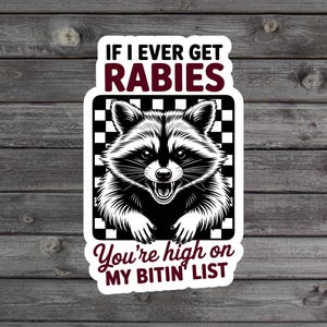 May include: A white sticker with a black and white raccoon illustration. The sticker has text that reads "IF I EVER GET RABIES You're high on MY BITIN' LIST" in a mix of fonts and colors. The background is a wooden surface.