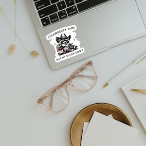 May include: A sticker with a cartoon raccoon wearing a cowboy hat and holding a jar of strawberry jam and a gun. The text on the sticker reads "Strawberry Jams But My Glock Don't".