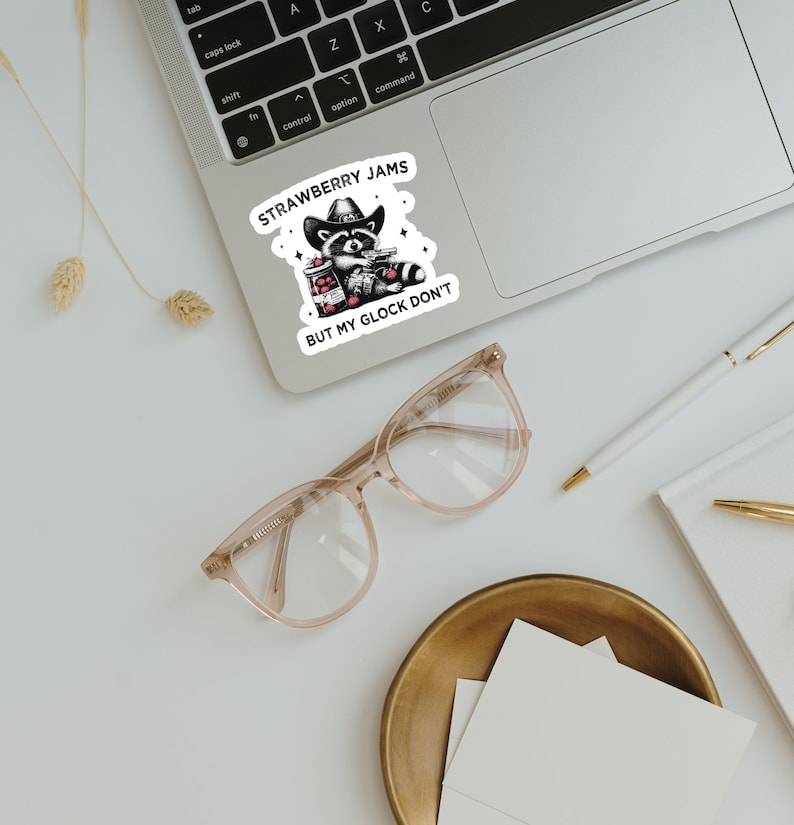 May include: A sticker with a cartoon raccoon wearing a cowboy hat and holding a jar of strawberry jam and a gun. The text on the sticker reads "Strawberry Jams But My Glock Don't".