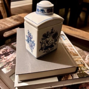 May include: A white ceramic tea caddy with a square shape and a rounded lid. The caddy is decorated with blue floral patterns and blue trim. It sits on top of a grey book.