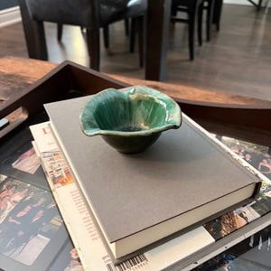 May include: A small, green ceramic bowl with a petal-like shape rests on two books on a wooden tray. The bowl has a glossy finish with darker green accents in the centre. The books are grey and white, with a photo-filled book visible below.