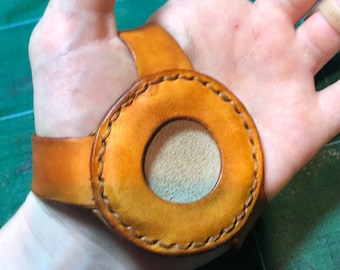 May include: A brown leather wrist strap with a circular cutout in the center. The strap is stitched with a brown thread and has a light brown leather lining.