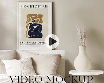 May include: A white frame with a poster inside. The poster has a beige background with a black and gold abstract figure. The text on the poster reads "MOCKTOPTRIE PSD SMART OBJECT MOCKTOPTRIE VIDEO FRAME MOCKUP SIMPLE AND EASY TO USE". The frame is on a white headboard with a white bedspread and pillows. A white side table with a vase of dried flowers is to the right of the frame. The text "VIDEO MOCKUP" is at the bottom of the image.