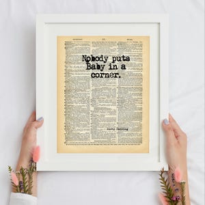 Peut inclure: Une impression encadrée présentant une page de dictionnaire vintage avec le texte noir "Nobody puts Baby in a corner." imprimé sur les colonnes. La page a un aspect papier vieilli beige avec des définitions et les mots "Dirty Dancing".