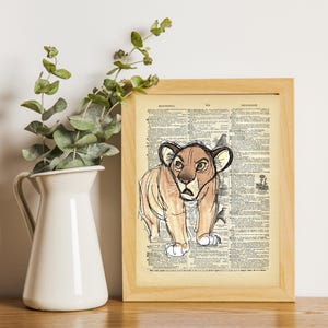 May include: A vintage dictionary page print featuring a hand-drawn illustration of a brown lion cub with a serious expression. The print is framed in a light wood frame.
