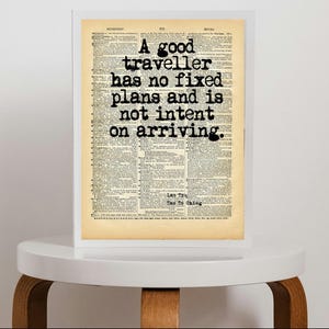 Op de afbeelding: Een rechthoekige wandkunstprint in een witte lijst met een achtergrond van een vintage woordenboekpagina en de zwarte tekst: A good traveller has no fixed plans and is not intent on arriving. Onderaan staat Lao Tzu, Tao Te Ching.
