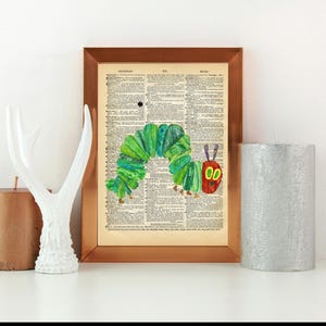 Könnte beinhalten: Ein gerahmter Druck einer grünen Raupe mit rotem Kopf und schwarzen Augen auf einer alten Wörterbuchseite. Der Rahmen ist kupferfarben.