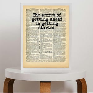 Puede incluir: Un marco blanco rectangular que contiene una página de diccionario antigua con papel color canela envejecido. El texto "The secret of getting ahead is getting started." está impreso en negrita con fuente de máquina de escribir sobre las columnas.