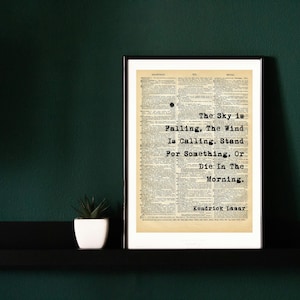 May include: A framed print of a vintage book page with a quote by Kendrick Lamar: "The Sky is Falling, The Wind Is Calling. Stand For Something, Or Die: In The Morning."