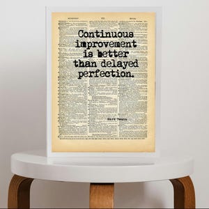 May include: A framed print featuring a vintage dictionary page background with the black text: Continuous improvement is better than delayed perfection. The quote is attributed to Mark Twain and is displayed in a white frame.
