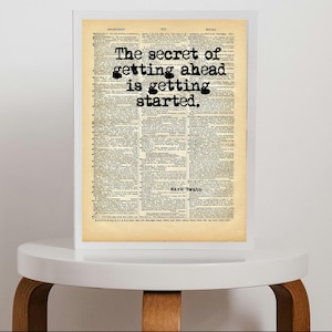 Pode incluir: Uma impressão emoldurada numa página de dicionário vintage com o texto preto em estilo de máquina de escrever: The secret of getting ahead is getting started. A citação é atribuída a Mark Twain sobre papel bege envelhecido.