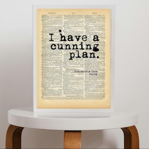 May include: A framed print featuring a vintage dictionary page with the black typewriter-style text "I have a cunning plan." and "Blackadder Goes Forth" printed over the columns of small, dense, aged beige paper text in a white frame.