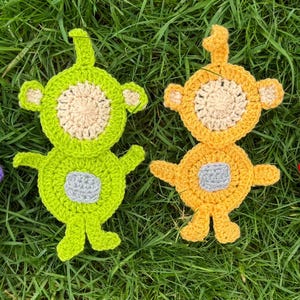 Pode incluir: Quatro personagens de crochê em forma de alienígenas, cada um com uma cor diferente: roxo, verde, amarelo e vermelho. Os personagens têm dois olhos e uma boca, e todos estão em pé em uma superfície verde gramada.