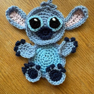 May include: A blue crocheted Stitch character from the Disney movie Lilo & Stitch. The character has large eyes, a blue nose, and pink ears. The character is sitting on a wooden surface.