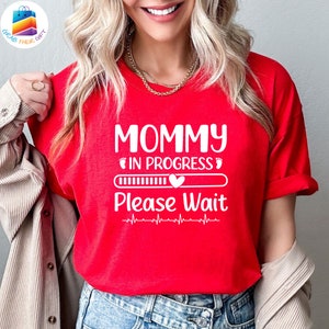 以下が含まれることがあります： 赤いTシャツに、白い文字で「Mommy in Progress」と書いてあり、プログレスバーとハートが付いています。「Please Wait」という文字がプログレスバーの下にあり、心臓の鼓動のグラフィックがあります。