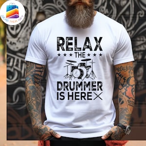 May include: White t-shirt with the text "RELAX THE DRUMMER IS HERE" in black. The design includes a drum set graphic. The shirt is short-sleeved.
