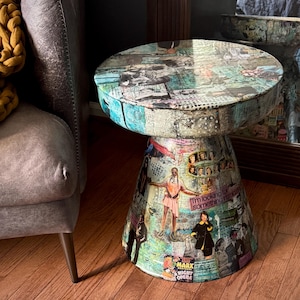 May include: A round side table with a tapered base, entirely covered in a colorful collage of vintage magazine clippings, photographs, and text. The surface features various human figures, graphic patterns, and the text "I'm looking for something intimidating" and "Marx Brothers A Night at the Opera."