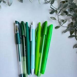 May include: A collection of six green and blue pens, including a clear pen with a silver tip, a dark green pen, a light green pen, a light blue pen, a dark green pen, and a bright green pen.