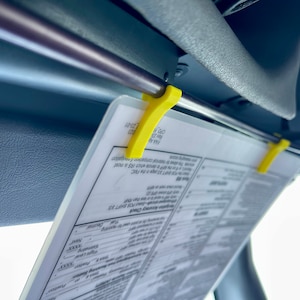 May include: A yellow plastic clip holds a white paper with the text "Auracy Check POS SHIFT 33 SHIFT 33 page in the FMC" and "FAA A 23-01" on it. The paper is attached to a gray car sun visor.