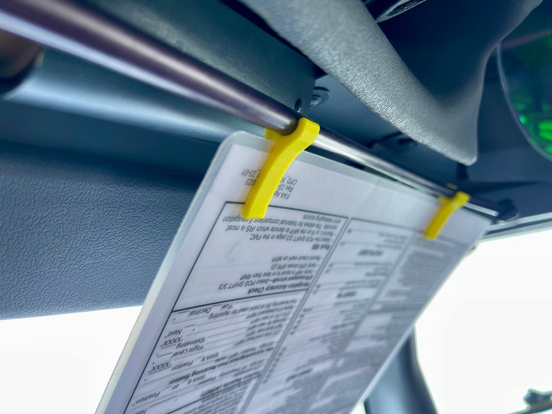 May include: A yellow plastic clip holds a white paper with the text "Auracy Check POS SHIFT 33 SHIFT 33 page in the FMC" and "FAA A 23-01" on it. The paper is attached to a gray car sun visor.