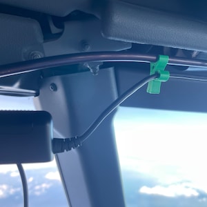 May include: A close-up of a black power adapter with a coiled cord, secured to a gray surface with a green cable clip. The cord is connected to a black cable running along a brown metal bar.