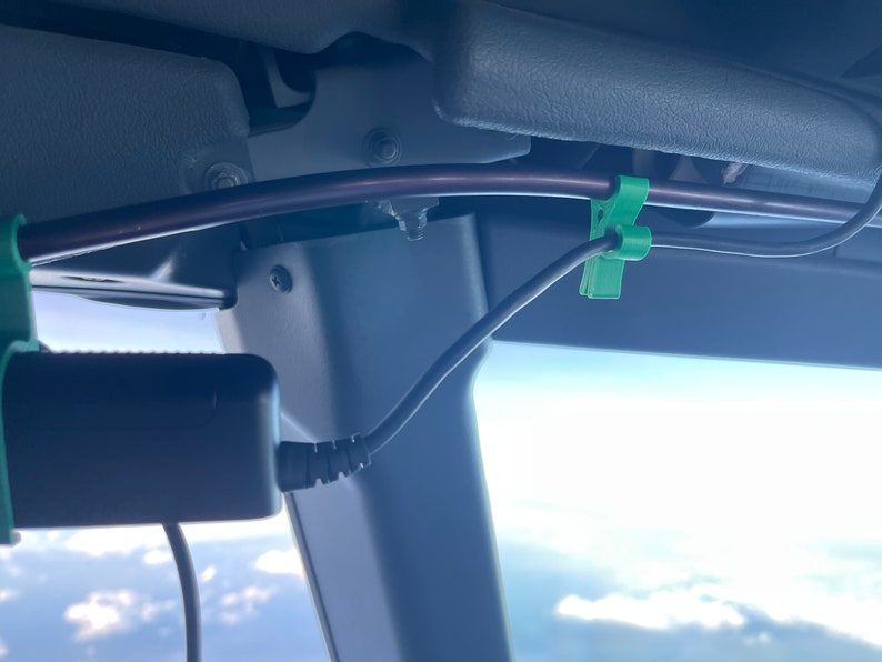May include: A close-up of a black power adapter with a coiled cord, secured to a gray surface with a green cable clip. The cord is connected to a black cable running along a brown metal bar.
