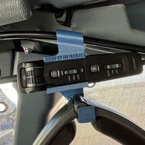 May include: A blue 3D printed holder with the text "MANLY ON THE PLANE.COM" attached to a black cord. The cord is connected to a black Bose noise-canceling headphone control unit with buttons for volume, ANC, high, med, low, mute, mix, and off.