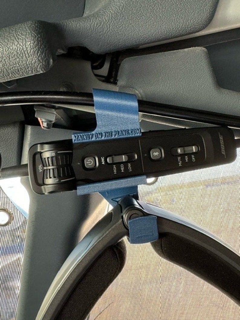 May include: A blue 3D printed holder with the text "MANLY ON THE PLANE.COM" attached to a black cord. The cord is connected to a black Bose noise-canceling headphone control unit with buttons for volume, ANC, high, med, low, mute, mix, and off.