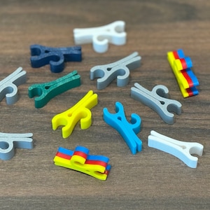 May include: A collection of colorful plastic clips in various shapes and sizes. The clips are arranged on a wooden surface. Some of the clips are in the shape of a clothespin, while others are more abstract. The clips are a mix of blue, green, yellow, and white.