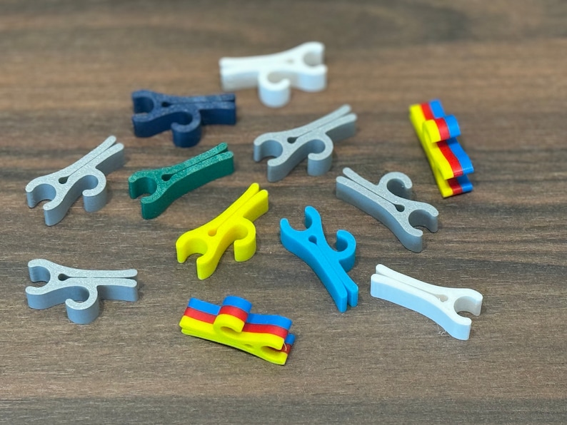 May include: A collection of colorful plastic clips in various shapes and sizes. The clips are arranged on a wooden surface. Some of the clips are in the shape of a clothespin, while others are more abstract. The clips are a mix of blue, green, yellow, and white.