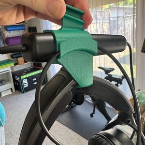 May include: A 3D printed green headphone holder with a black headphone attached. The holder has a curved design and a small lip on the top.