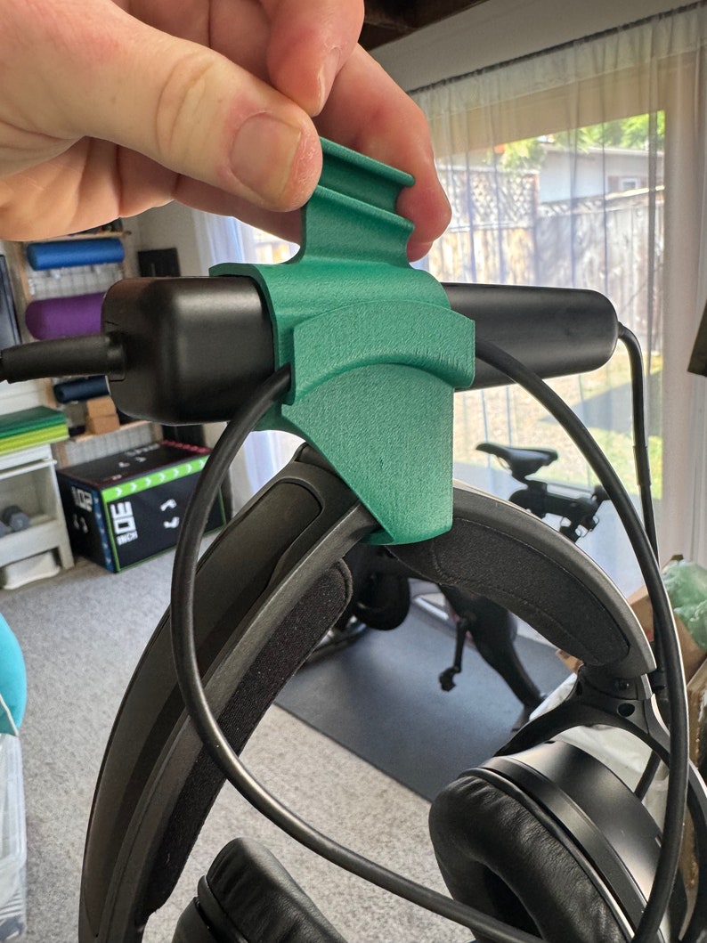 May include: A 3D printed green headphone holder with a black headphone attached. The holder has a curved design and a small lip on the top.