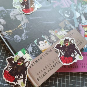 Puede incluir: Primer plano de una novela gráfica de Batman con un marcapáginas rojo y negro que presenta un personaje con máscara y una cadena. El marcapáginas tiene el texto "The Killing Joke". La portada del libro presenta el texto "Batman" y "Ciudad Roja" en español.